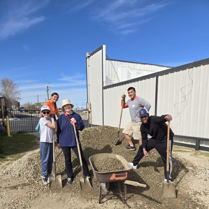 A busy kitchen scene with volunteers preparing meals, assembling food containers, and various kitchen supplies visible in the background.